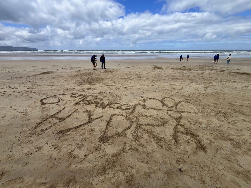 “HYDRA” and a fleur-de-lis symbol drawn in the sand during a beach activity.